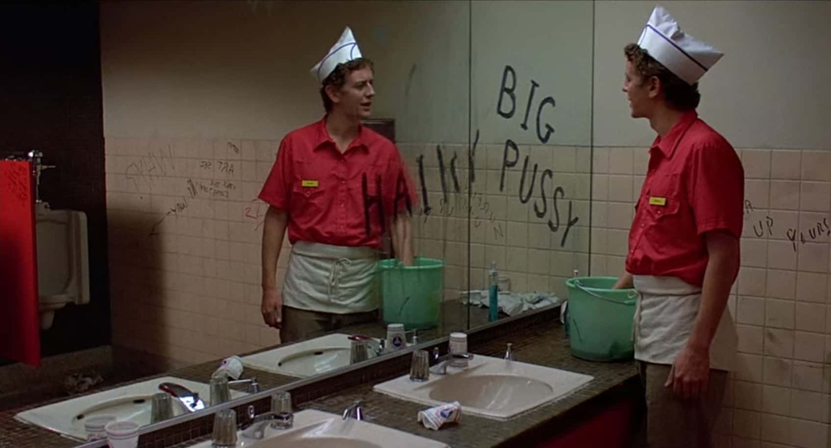 Brad Hamilton stands in a restroom, facing a mirror with crude graffiti and cleaning supplies nearby