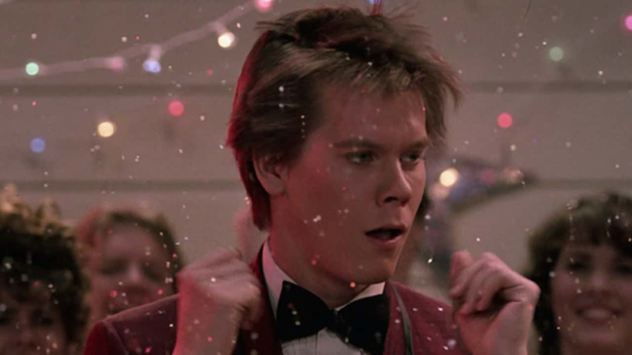 Ren McCormack dances awkwardly in a festive setting from Footloose, with onlookers in the background